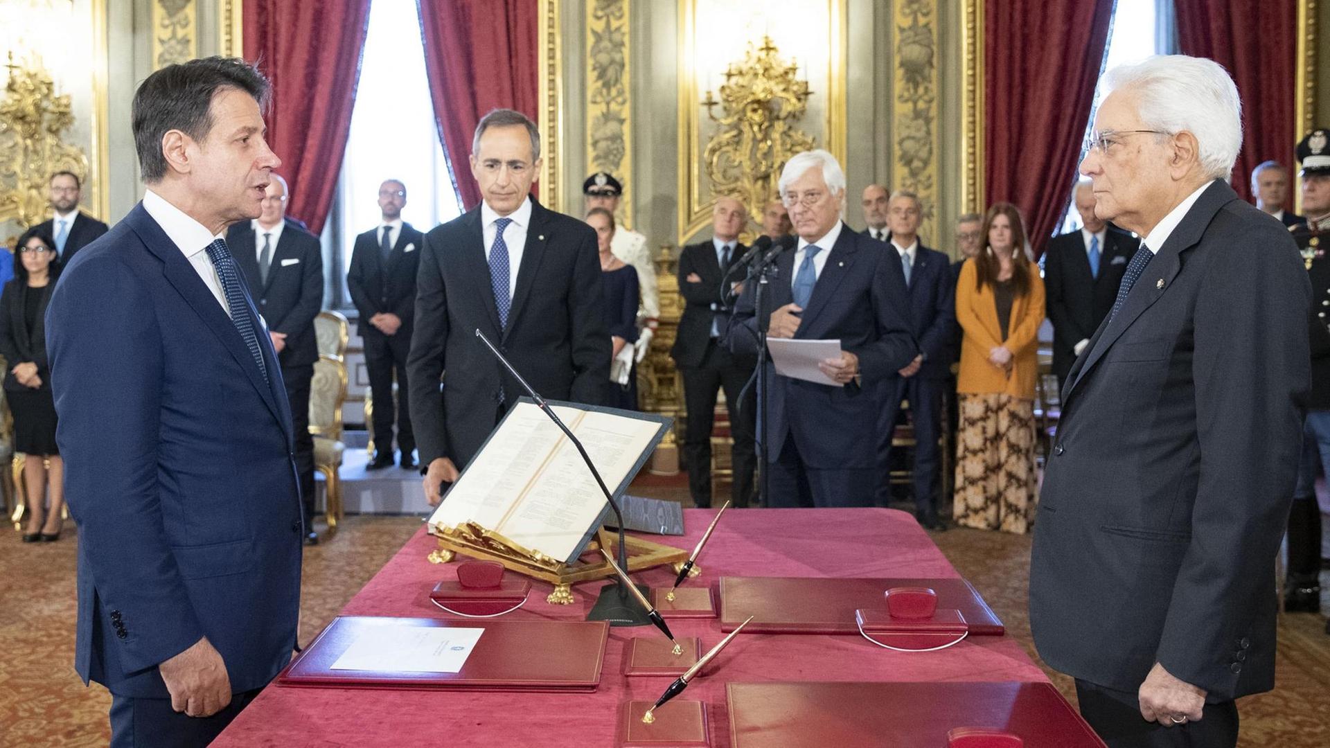 Conte und Mattarelle stehen sich an einem roten Tisch gegenüber. Im Hintergrund stehen andere Personen. Conte und Mattarelle stehen sich an einem roten Tisch gegenüber. Im Hintergrund stehen andere Personen.