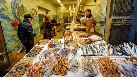 Das Bild zeigt ein Fischgeschäft in Bologna, Italien. In der Auslage liegt der Frischfisch, ein Verkäufer reicht einer Kundin eine Tüte.