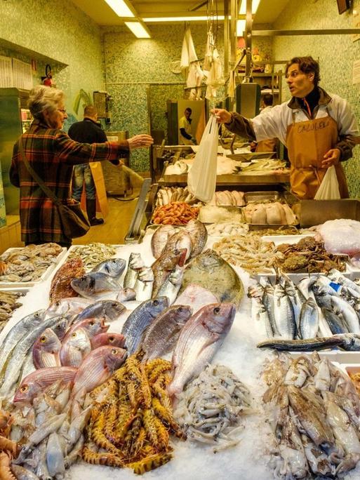 Das Bild zeigt ein Fischgeschäft in Bologna, Italien. In der Auslage liegt der Frischfisch, ein Verkäufer reicht einer Kundin eine Tüte.
