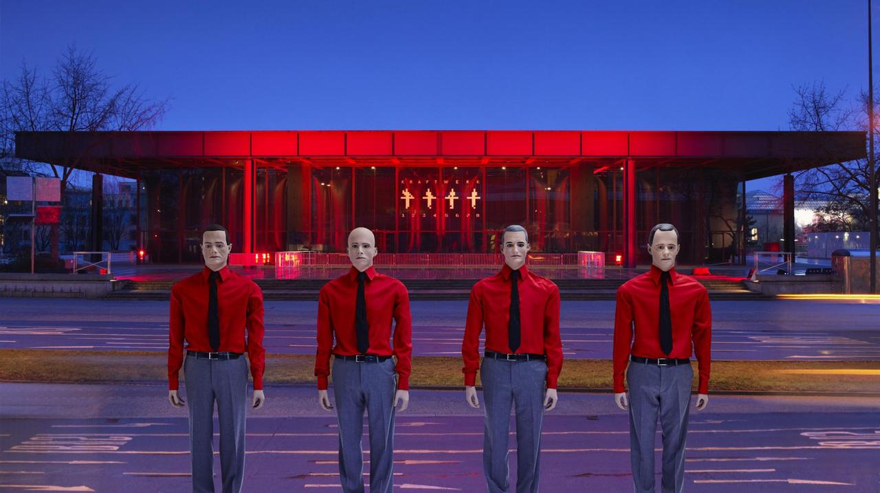 Die Musiker von Kraftwerk stehen mit roten Hemden nebeneinander, im Hintergrund ein rot angeleuchtetes Gebäude.
