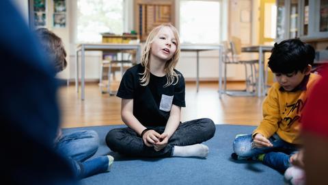 Ein Mädchen sitzt in einem Schulraum auf dem Boden. Links und rechts sitzen weitere Kinder.