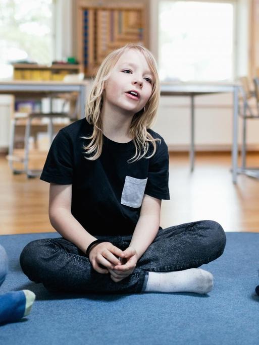 Ein Mädchen sitzt in einem Schulraum auf dem Boden. Links und rechts sitzen weitere Kinder.