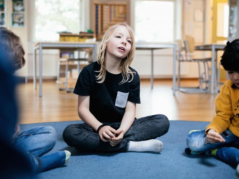 Ein Mädchen sitzt in einem Schulraum auf dem Boden. Links und rechts sitzen weitere Kinder.