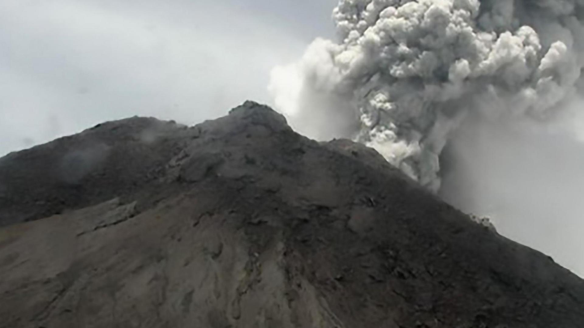 Blick aus weiter Ferne auf den schwarzen Vulkan, aus dem grauer Rauch aufsteigt.