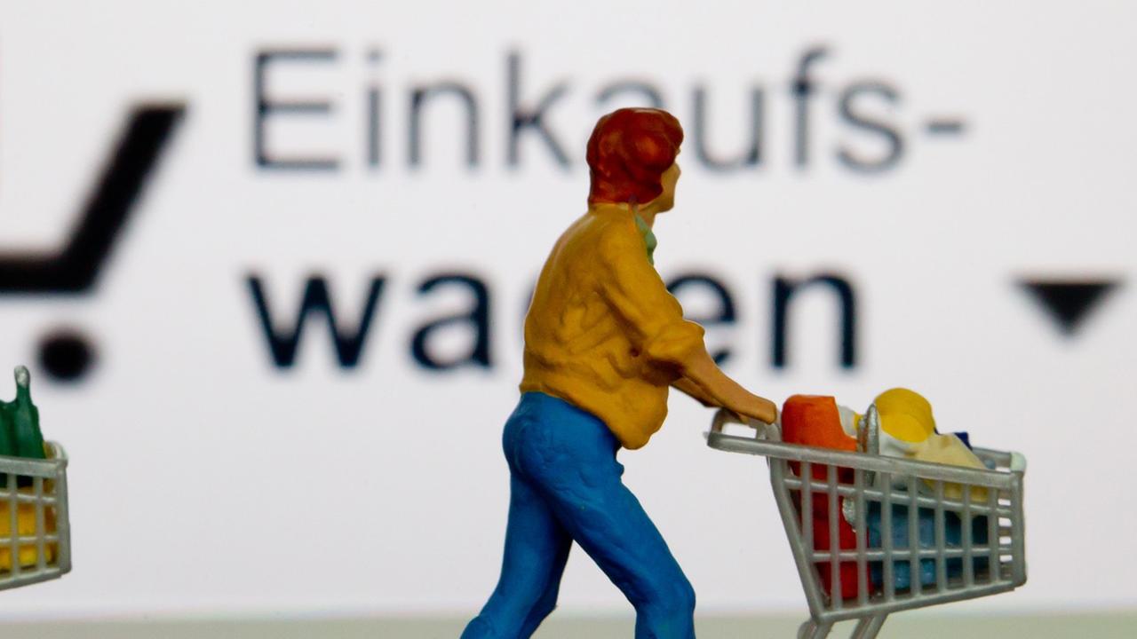 Zwei kleine Plastikfiguren mit Einkaufswagen stehen vor einem iPhone, auf dem der virtuelle Einkaufswagen dargestellt ist.