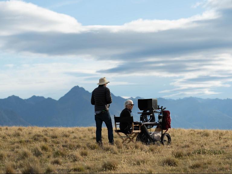 Auf einem Feld sitzt die Regisseurin Jane Campion auf einem Klappstuhl, vor ihr ein Monitor, im Hintergrund die Berge. Neben ihr steht ein Mann mit Cowboyhut