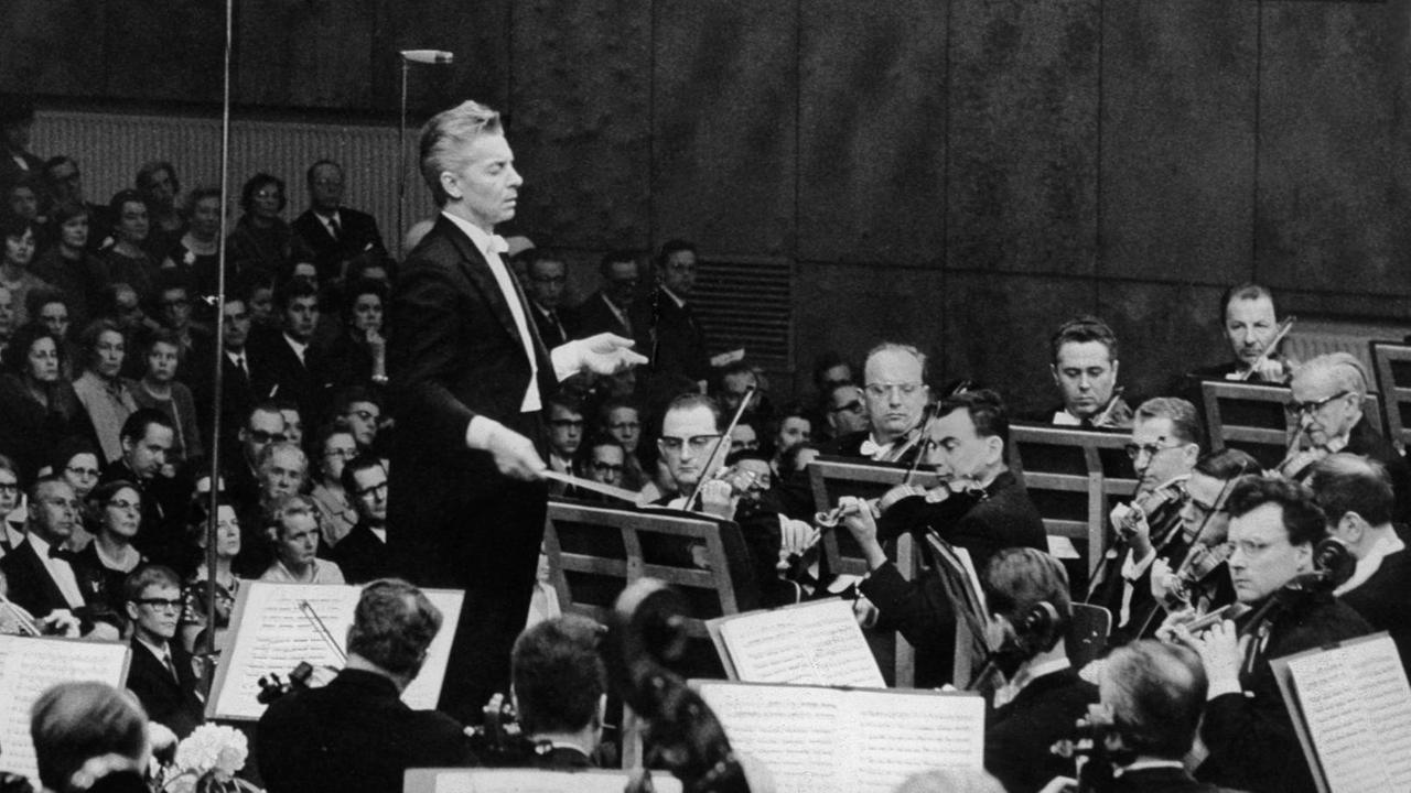 Schwarz-weiß Foto, seitliche Aufnahme. Der Dirigent, Herbert von Karajan, steht vor dem Orchester, er hält den Taktstock in der rechten Hand, er trägt einen Frack mit weißem Hemd und weißter Fliege. Rechts spielen die Musiker, der Publikumsraum ist gefüllt.