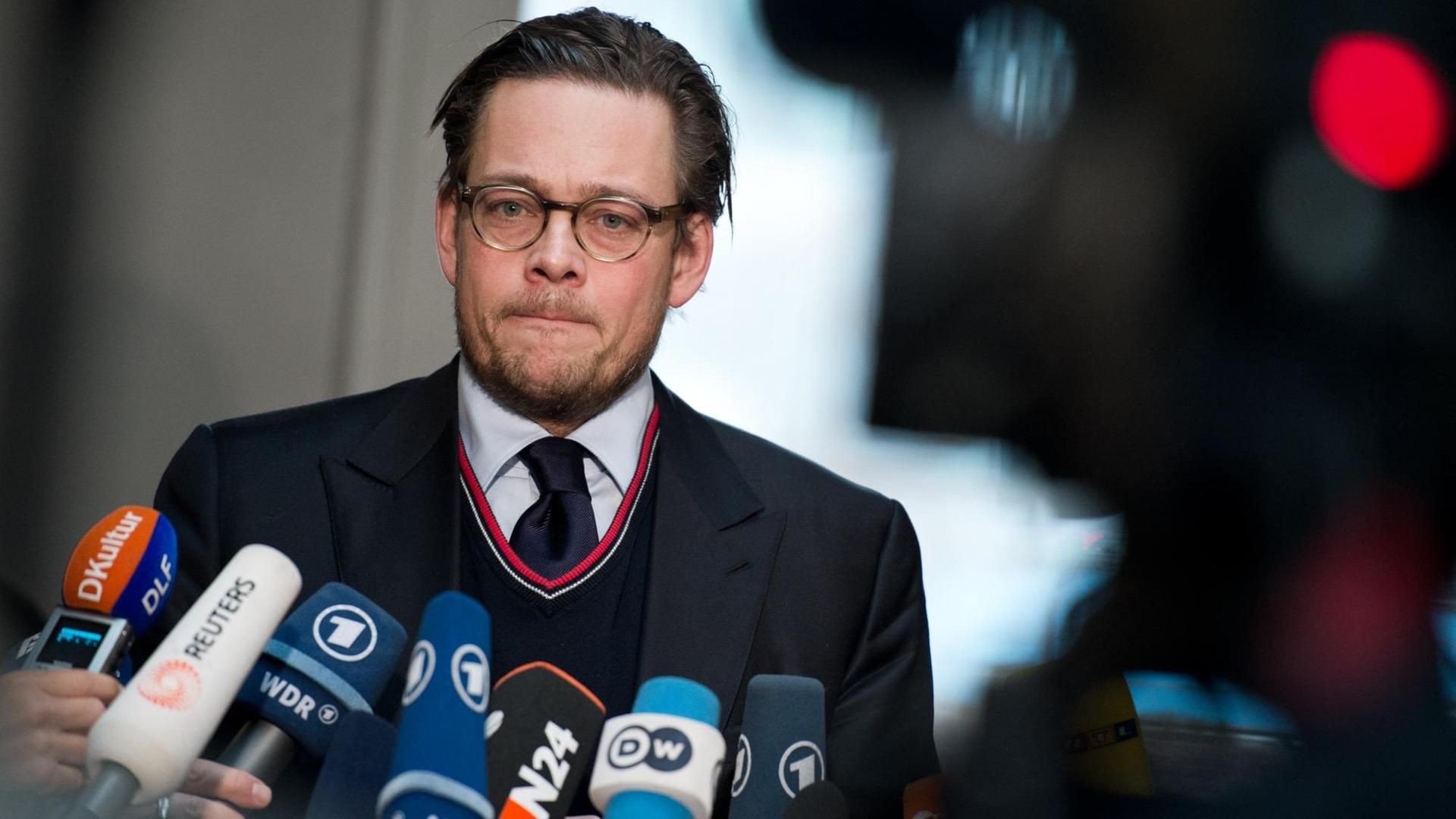 Ein Mann mit kurzen Haaren und Brille presst seine Lippen aufeinander – ihm sind viele Mikrofone und eine Kamera entgegen gestreckt.