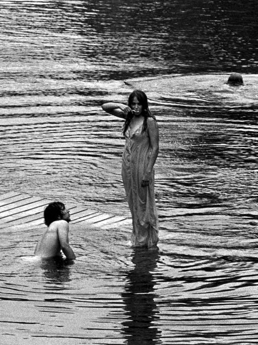 Schwarz-Weiß-Fotografie einer Frau, die im nassen Kleid auf dem Wasser zu stehen scheint, neben ihr schwimmen zwei Männer im See. 