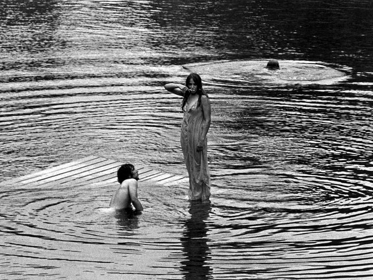 Schwarz-Weiß-Fotografie einer Frau, die im nassen Kleid auf dem Wasser zu stehen scheint, neben ihr schwimmen zwei Männer im See. 