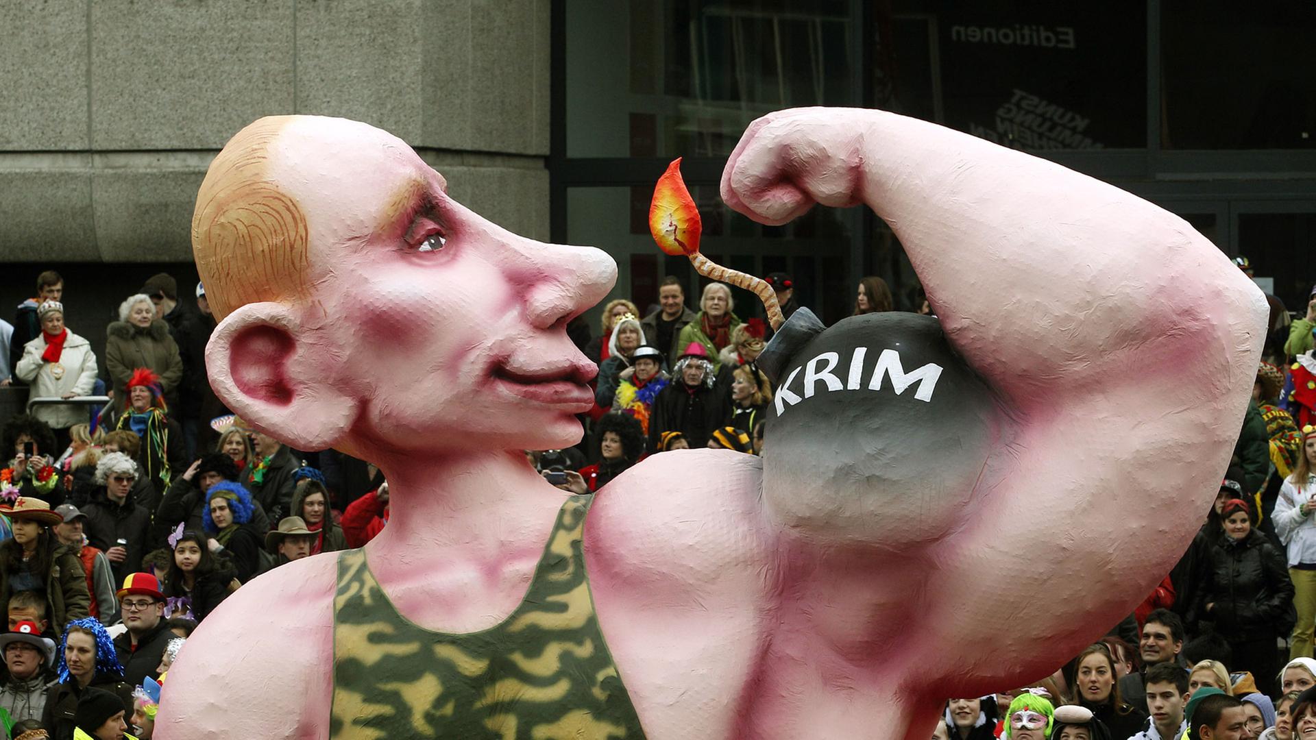 Ein Motivwagen auf dem Düsseldorfer Karnevalszug zeigt einen übergroßen Wladimir Putin in einem Camouflage-Unterhemd , der seinen Bizeps zeigt, auf dem "Krim" steht.