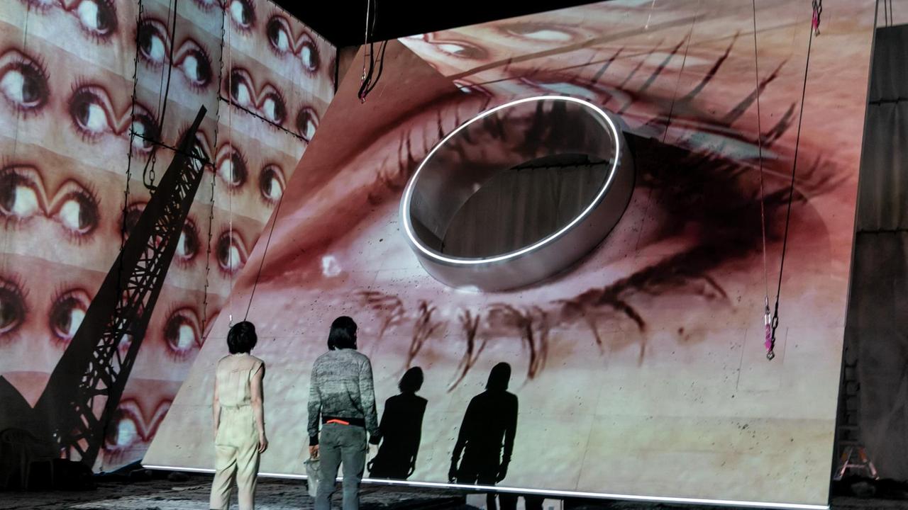 Das Foto aus dem Theaterstück "Evolution" zeigt ein überdimensioniertes Auge vor dem zwei Menschen, mit dem Rücken zum Publikum, stehen
