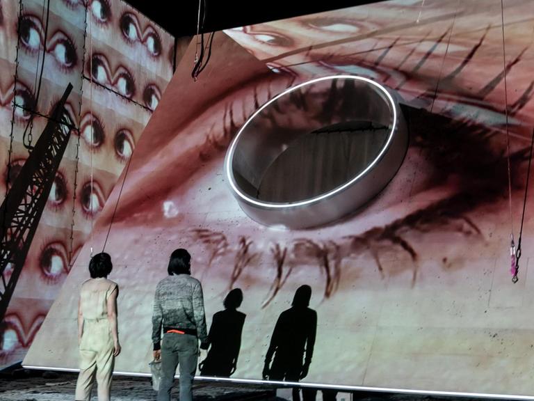 Das Foto aus dem Theaterstück "Evolution" zeigt ein überdimensioniertes Auge vor dem zwei Menschen, mit dem Rücken zum Publikum, stehen