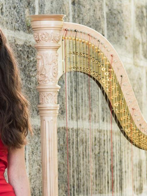 Die Musikerin steht in rotem Kleid neben ihrer Harfe, beide vor einer massiven, steinernen Mauer.