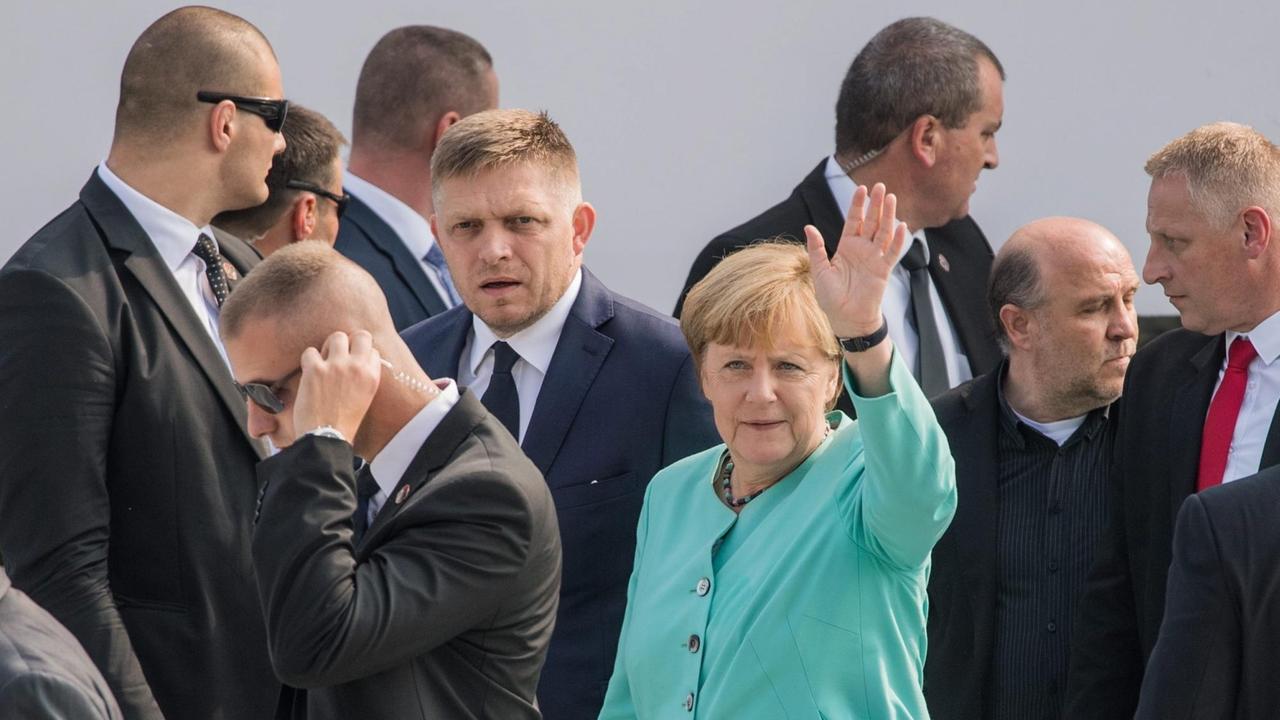 Merkel im grünen Kostum winkt mit der Hand, links neben ihr Fico. Beide sind umgeben von den Sicherheitsleuten. 