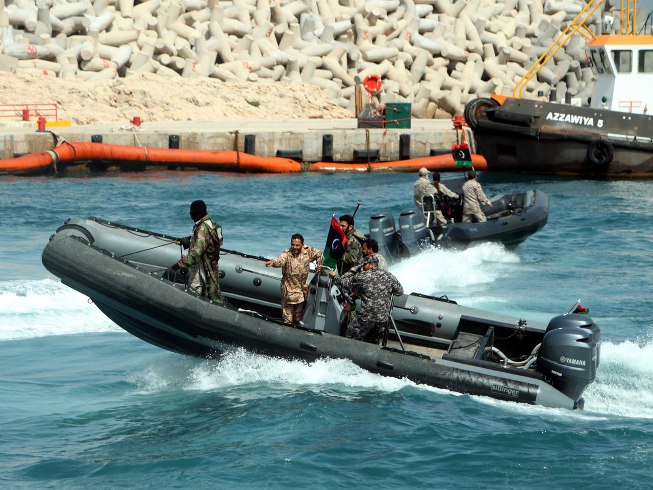 Ein flaches Motorboot mit mehreren Menschen an Bord fährt am Hafen übers Wasser, im Hintergrund der Kai und ein Fischerboot.