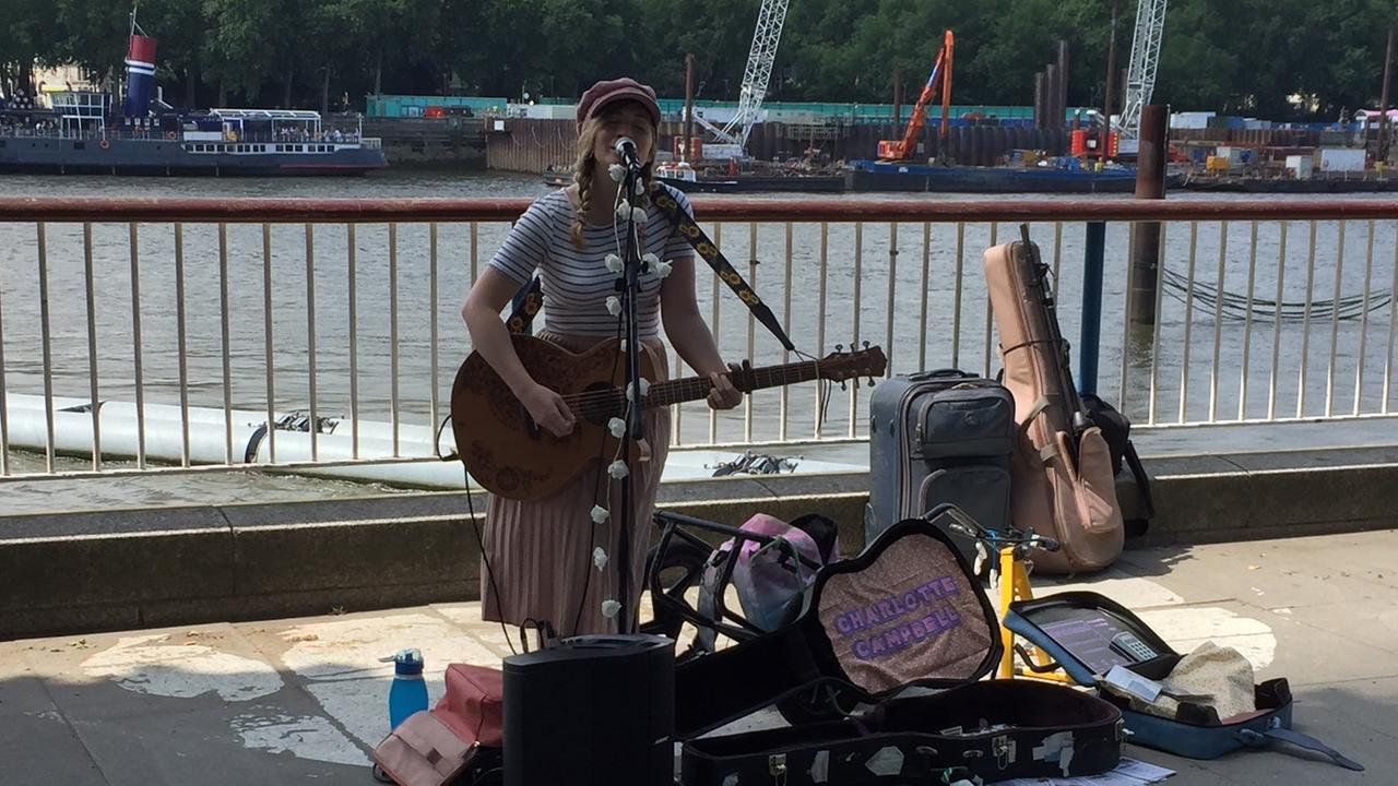 Strassenmusikerin Charlotte Campbell steht mit Gitarre und aufgeklappten Gitarrenkoffer für Geldspenden an der Themse in London