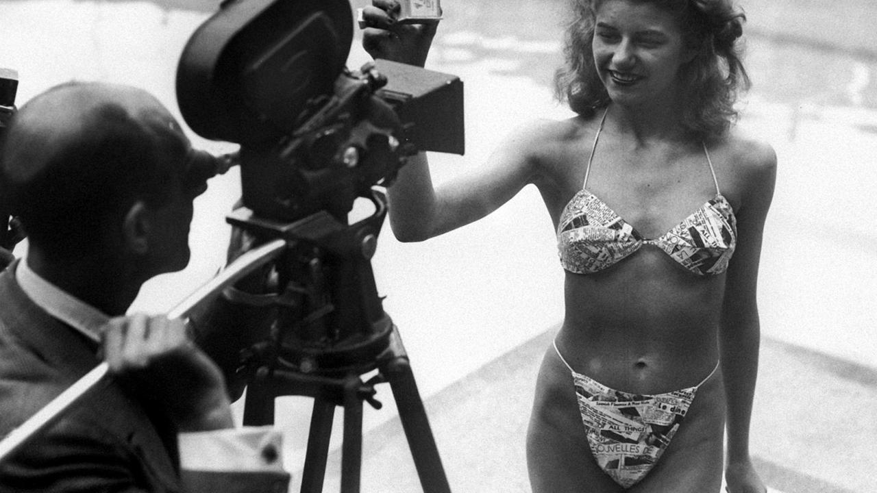 Ein Schwarz-Weiß-Bild zeigt einen Fotografen, der durch eine Kamera auf das Model Micheline Bernardini blickt, die am 5. Juli 1946 in einem Schwimmbad den ersten Bikini vorführt