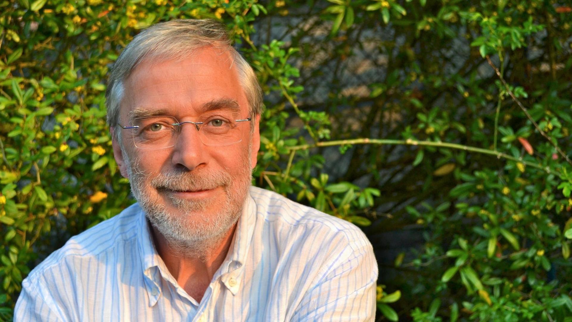 Der Neurobiologe Gerald Hüther, fotografiert in einem hell gestreifen Hemd vor einer grünen Hecke
