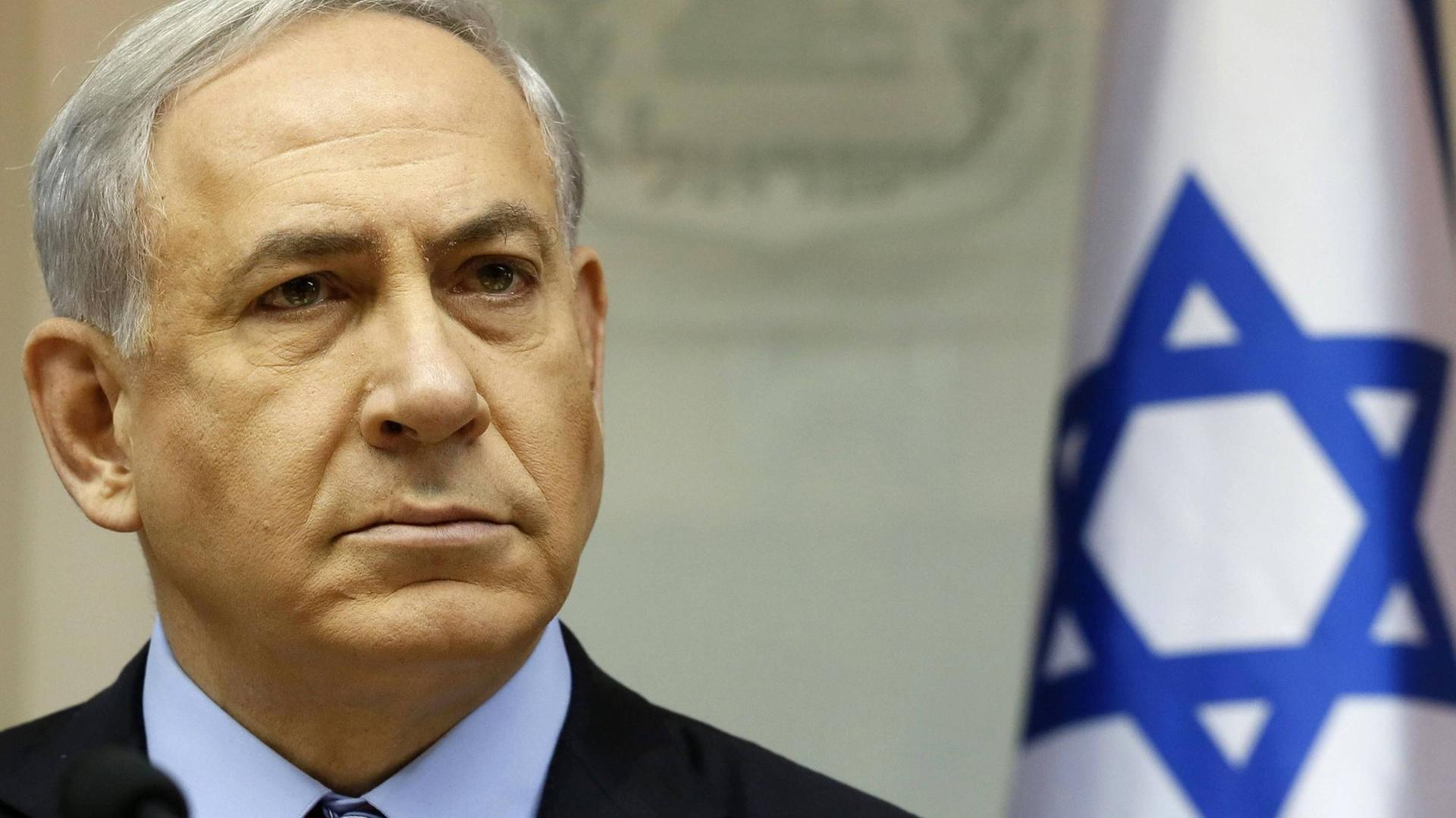 Benjamin Netanjahu steht mit ernstem Gesicht neben einer Israel-Fahne
