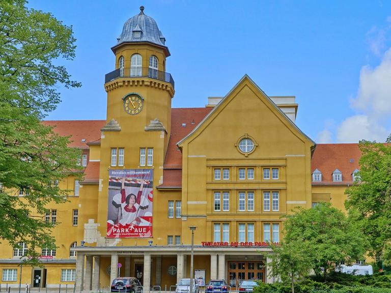 Außenansicht des Theaters an der Parkaue in Berlin-Lichtenberg. Ein großes Gebäude mit gelber Fassade, einem Spitzdach und einem kleinen Türmchen.
