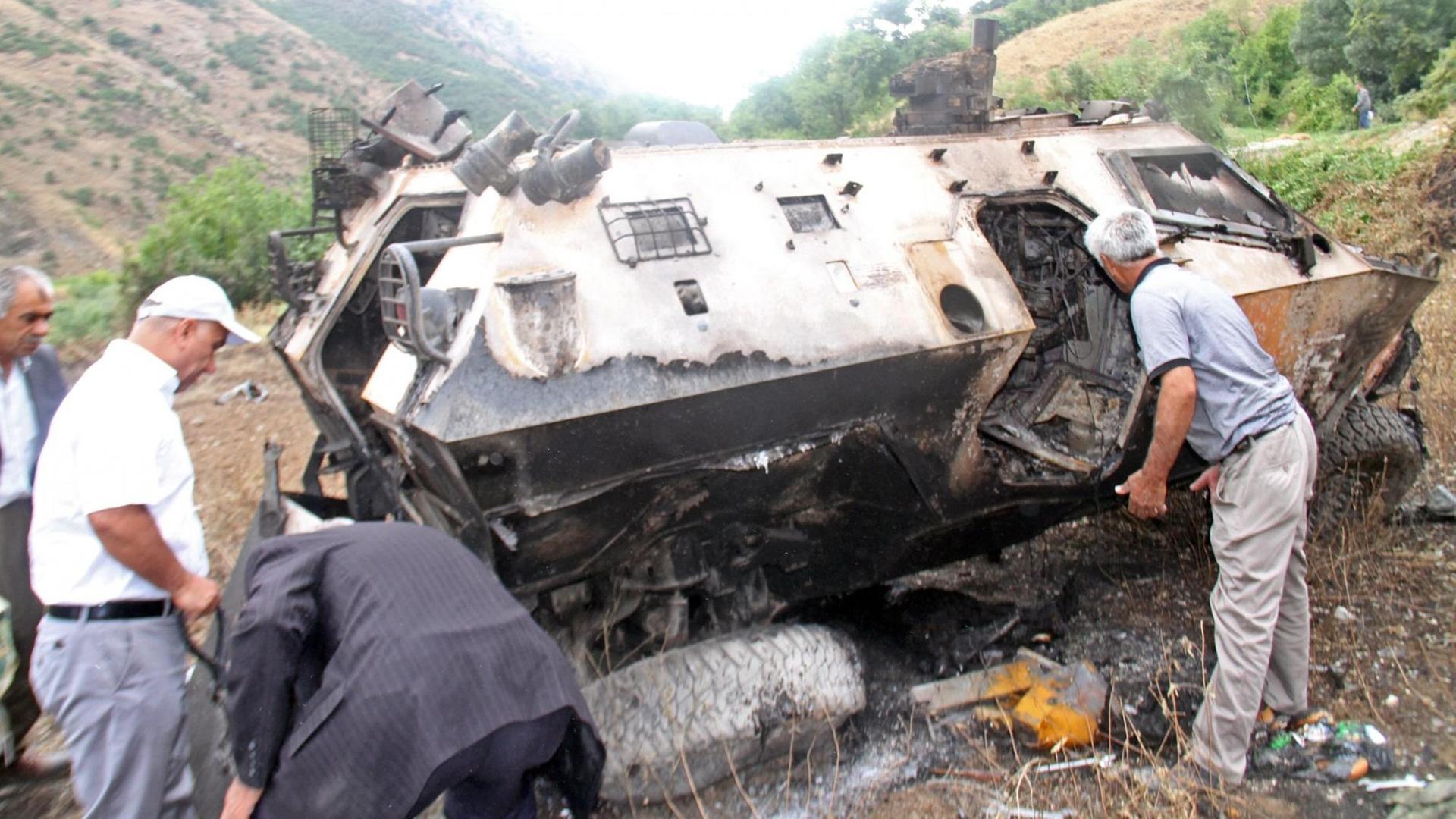 Ein ausgebranntes Fahrzeug der türkischen Armee liegt in einer gebirgigen Gegend. Mehrere Kurden stehen daneben und untersuchen das Wrack.