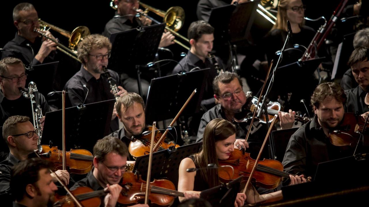 Blick in Orchesterreihen, in denen Musikerinnen und Musiker mit Geigen, Klarinetten und Posaunen zu sehen sind.