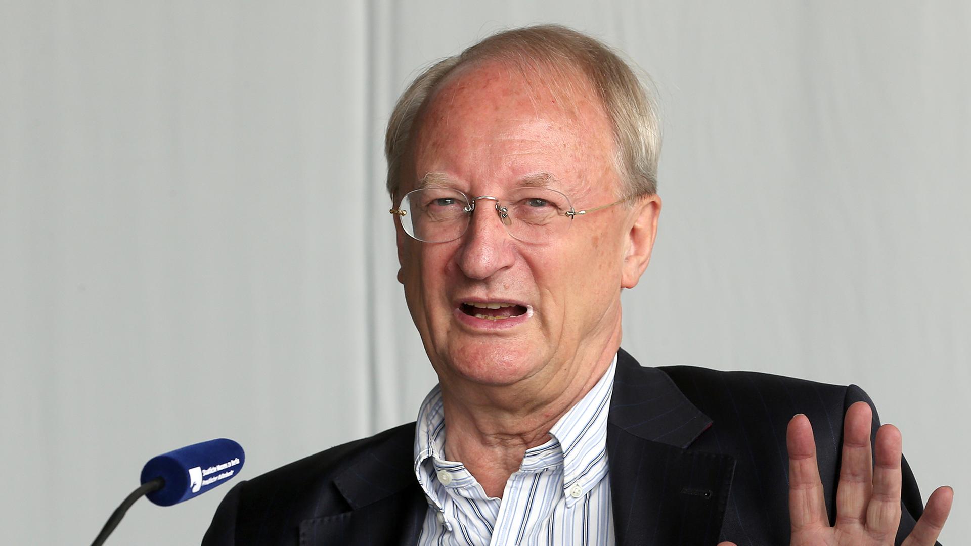 Klaus Staeck sitzt vor einem Mikrofon und hebt die Hand
