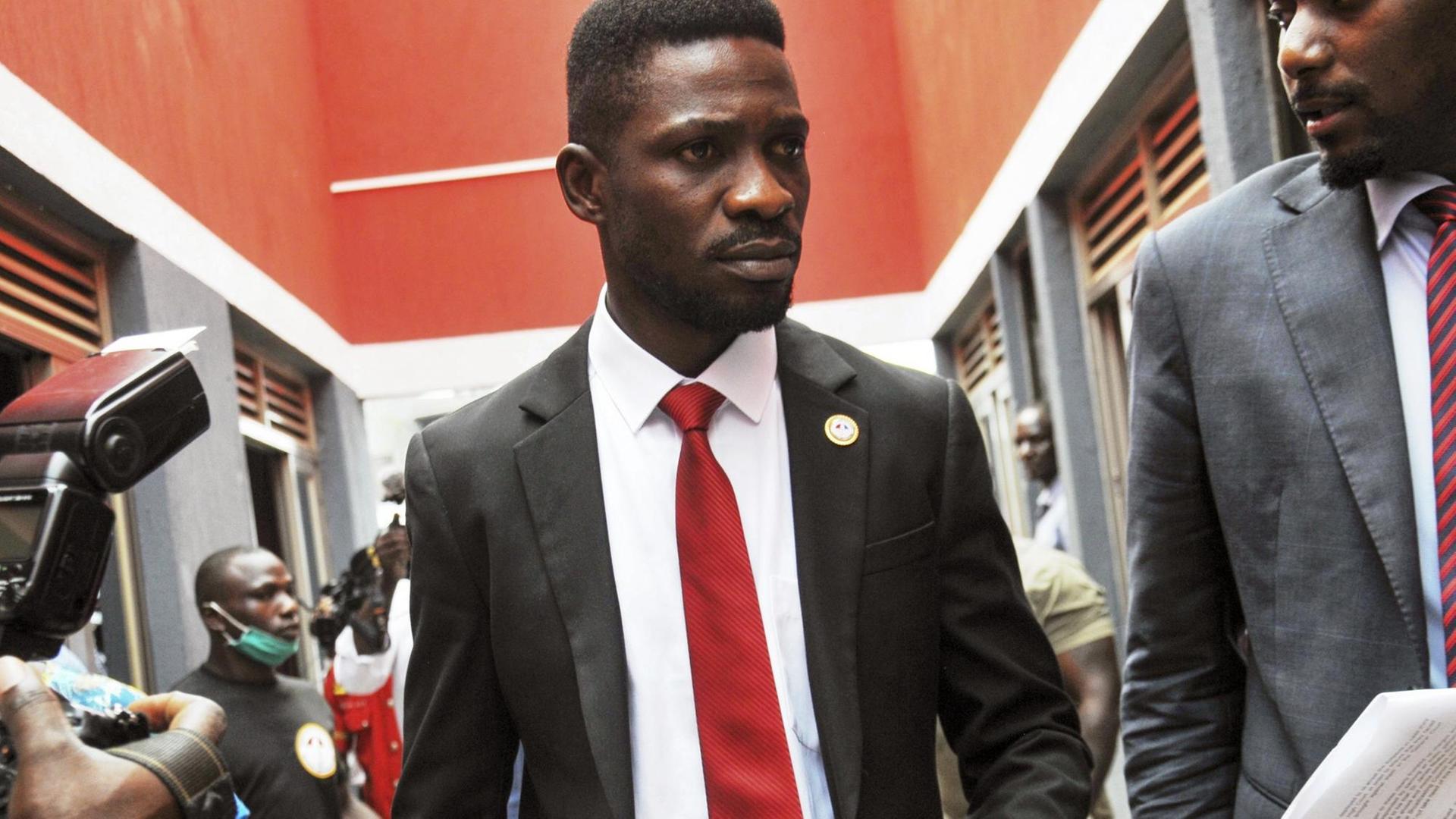 Bobi Wine im Anzug mit roter Krawatte geht durch einen Flur. Daneben und dahinter weitere Personen.