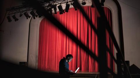 Ein Regisseur schaut im Zuschauerraum eines Theaters auf einen Laptop. Im Hintergrund: die Bühne mit geschlossenem Vorhang.