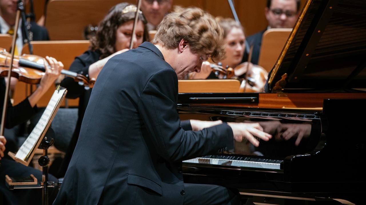 Der Pianist Jan Lisiecki sitzt auf einer Bühne und spielt Klavier. Im Hintergrund das Orpheus Chamber Orchestra.