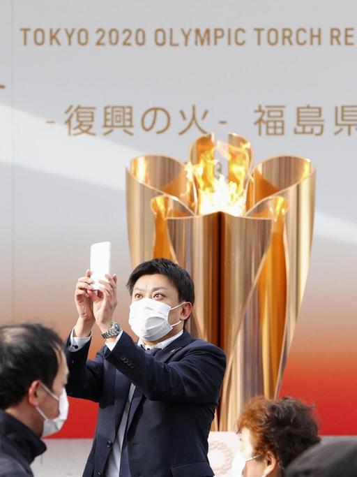 24.03.2020, Japan, Fukushima: Ein Besucher mit Mundschutz macht ein Selfie mit der olympischen Flamme in Fukushima. Die Flamme tourt durch die von der Erdbeben-Tsunami-Katastrophe im März 2011 schwer getroffenen Gebiete im Nordosten Japans, bevor der Fackellauf in der Präfektur Fukushima am 26. März beginnt. Foto: kyodo/dpa +++ dpa-Bildfunk +++