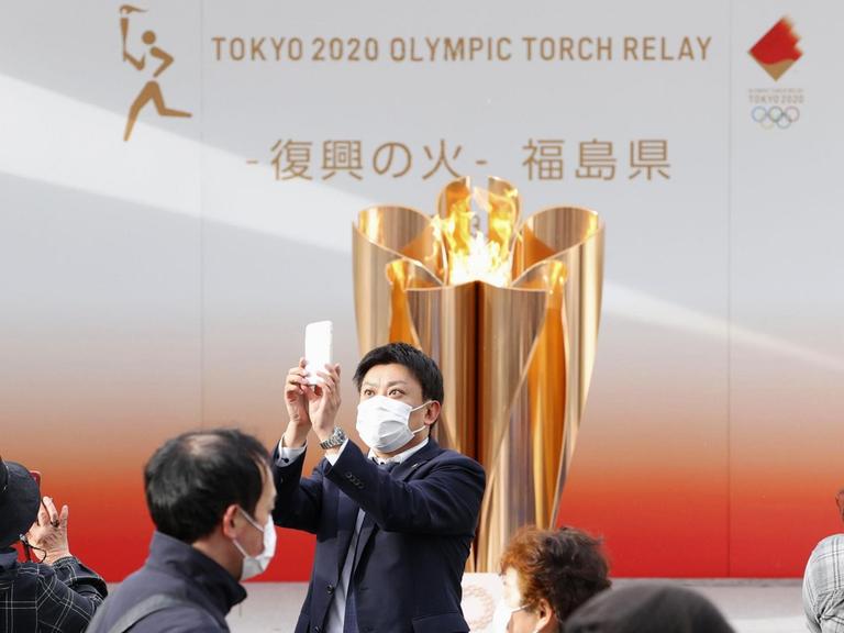 24.03.2020, Japan, Fukushima: Ein Besucher mit Mundschutz macht ein Selfie mit der olympischen Flamme in Fukushima. Die Flamme tourt durch die von der Erdbeben-Tsunami-Katastrophe im März 2011 schwer getroffenen Gebiete im Nordosten Japans, bevor der Fackellauf in der Präfektur Fukushima am 26. März beginnt. Foto: kyodo/dpa +++ dpa-Bildfunk +++