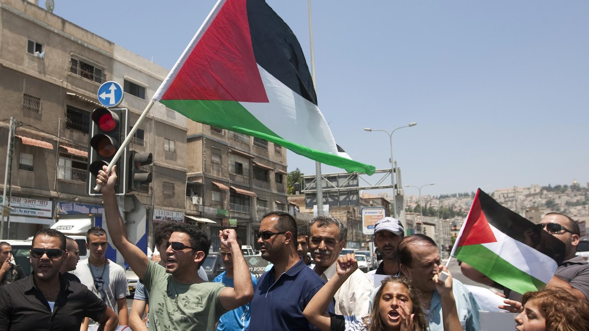 Menschen auf einer Straße vor Häusern, halten die palästinensische Flagge hoch, Arme in der Luft, rufen.
