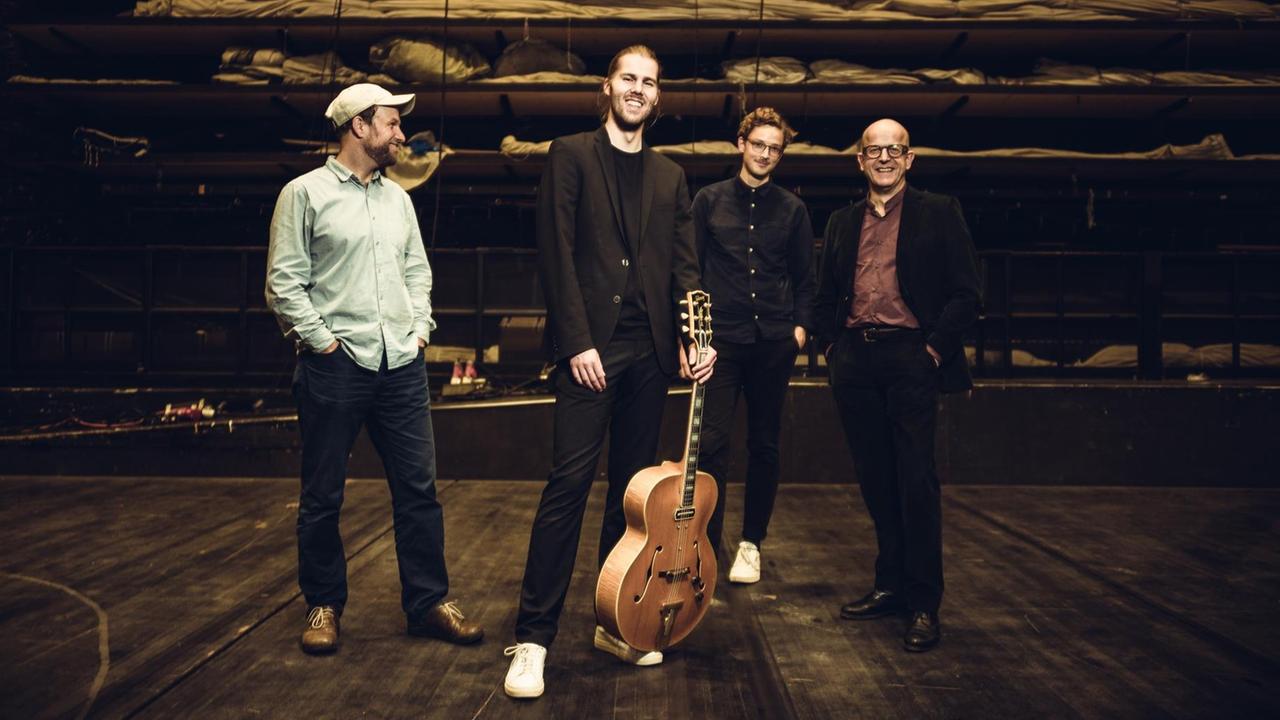 Auf einer großen, dunklen Bühne stehen vier Männer freundlich lachend nebeneinander. Der zweite von links hält eine auf seinem Fuß stehende Gitarre am Griffbrett fest.