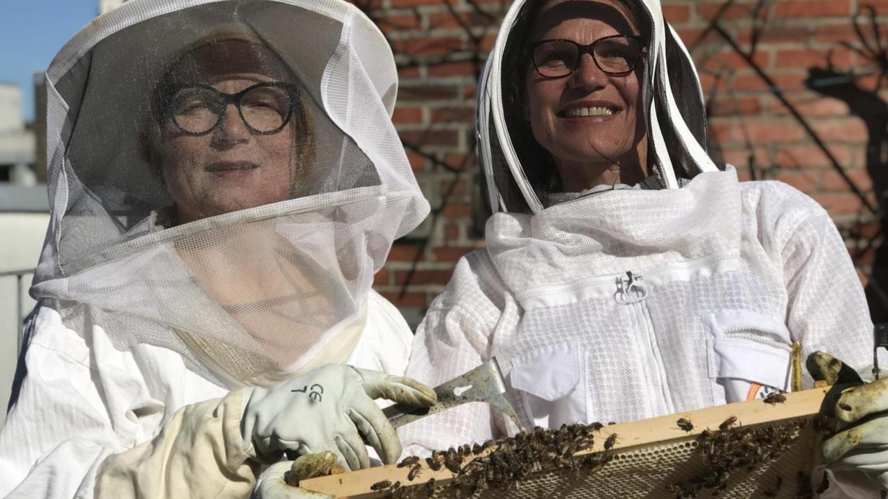 Das Bild zeigt die beiden Imkerinnen Iris Pinkepank und Stephanie Breil vom Projekt "HonigConnection" im Imkerschutzanzug mit einem Wabenrähmchen voller Bienen in den Händen.