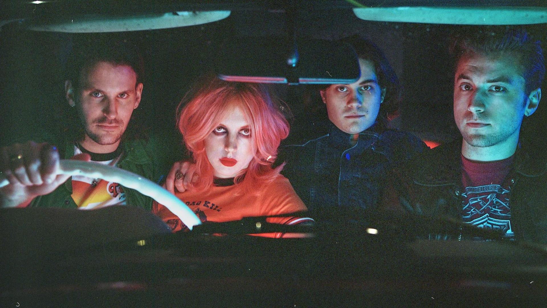 Drei Männer und eine Frau sitzen in einem Rechtslenker-Auto und blicken direkt in die Kamera. Vorne sitzen drei Personen, eine auf der Rückbank.