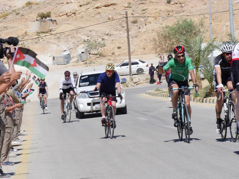 Kinder mit jordanischen Fahnen stehen am Rande einer Radstrecke, Rennradfahrer fahren vorbei.