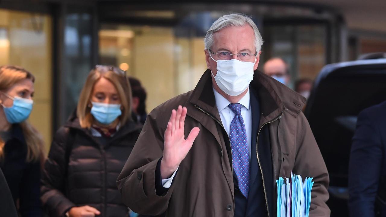 Barnier im braunen Mantel und mit Maske winkt in die Kamera. Er trägt einen Stapel Dokumente unter dem Arm. Hinter ihm unscharf weitere Personen.