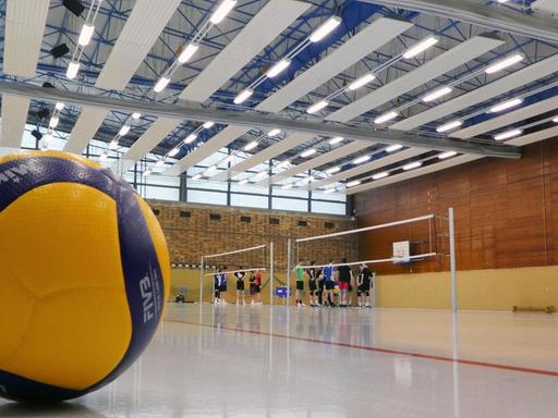 Sporthalle mit Spielern, die sich am Spielfeld bespechen. Im Vordergrund ist ein gelb-blauer Volleyball zu sehen.