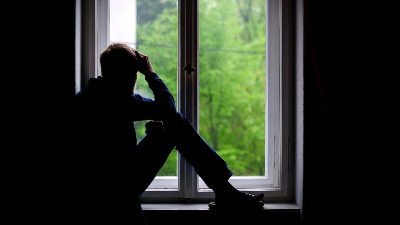 Ein Mann sitzt in einem dunklen Raum an einem Fenster und schaut hinaus