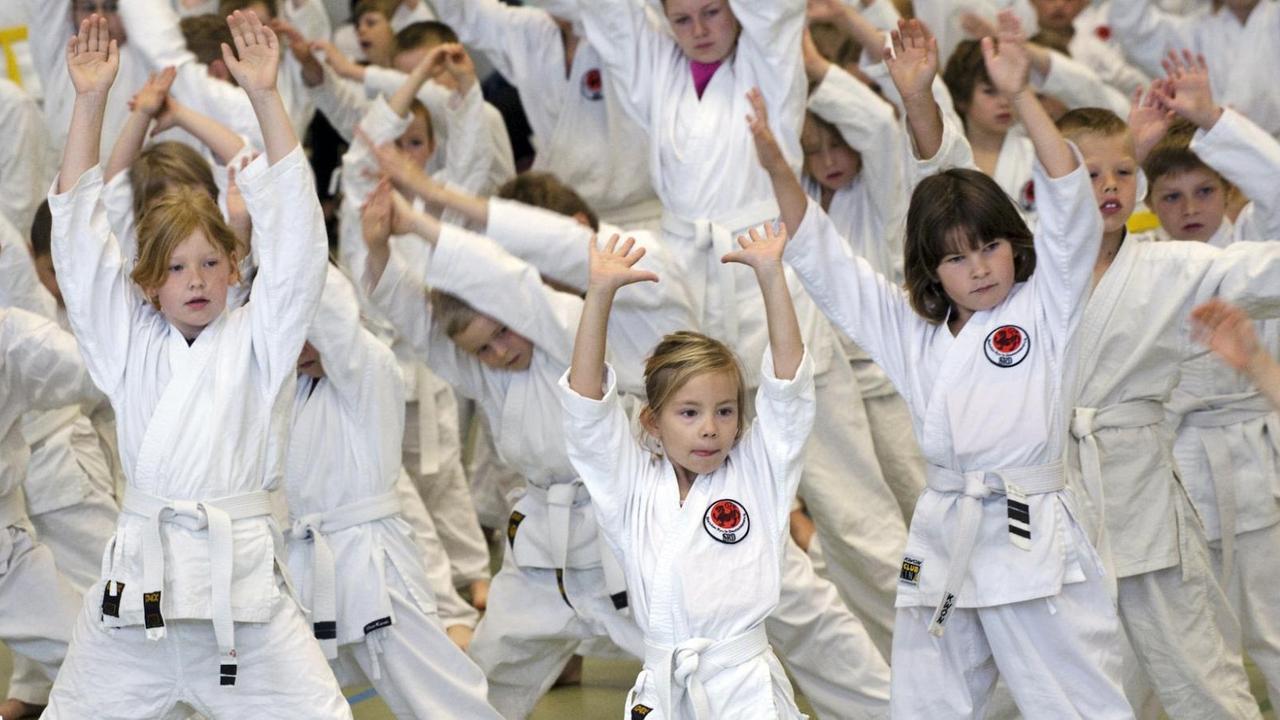 Junge Karate-Sportler in weißer Trainingskleidung üben eine Bewegung mit erhobenen Armen aus.