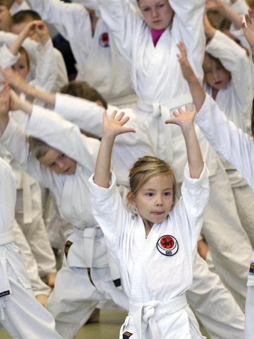 Junge Karate-Sportler in weißer Trainingskleidung üben eine Bewegung mit erhobenen Armen aus.
