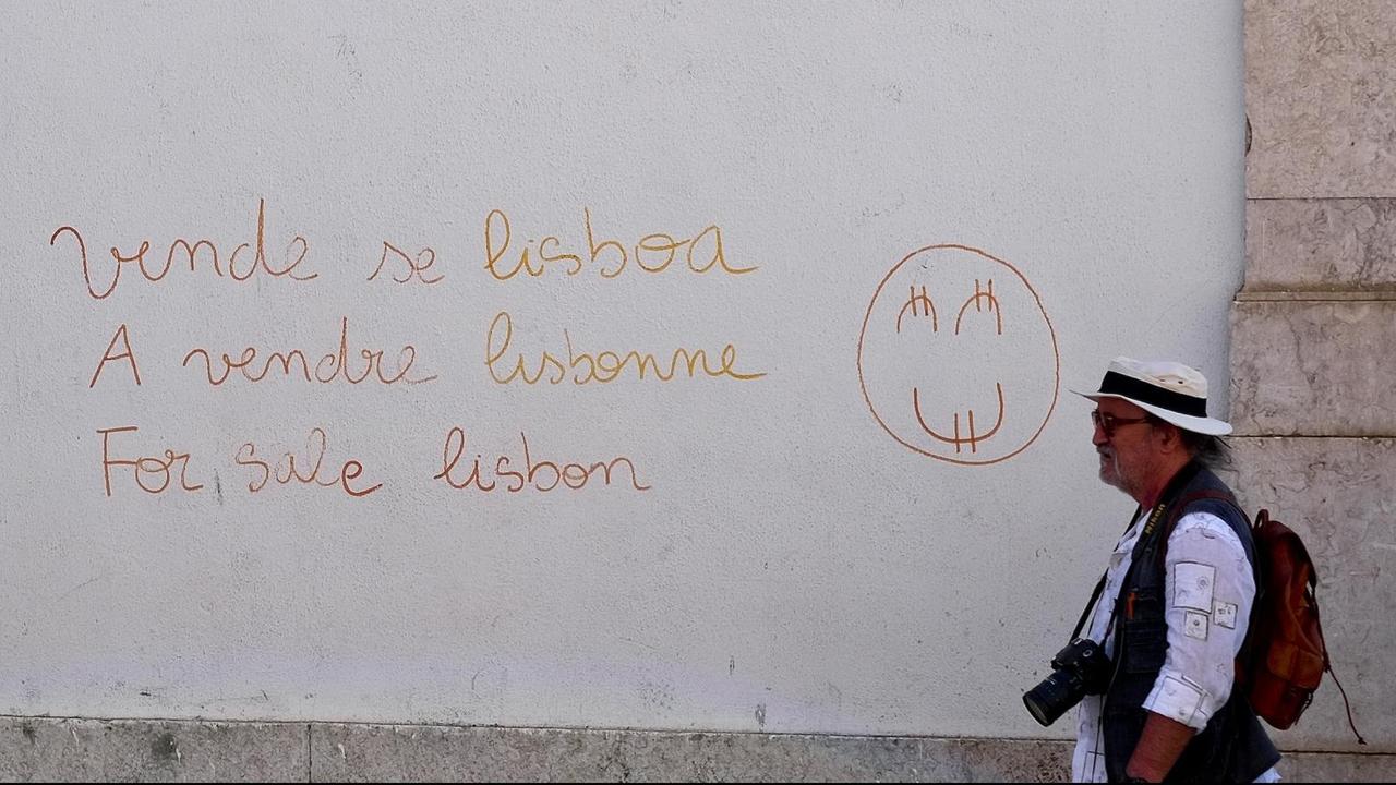 Touristen gehen in der Altstadt ovn Lissabon an einer Wand vorbei, auf der in mehreren Sprachen "Lissabon zu verkaufen" steht