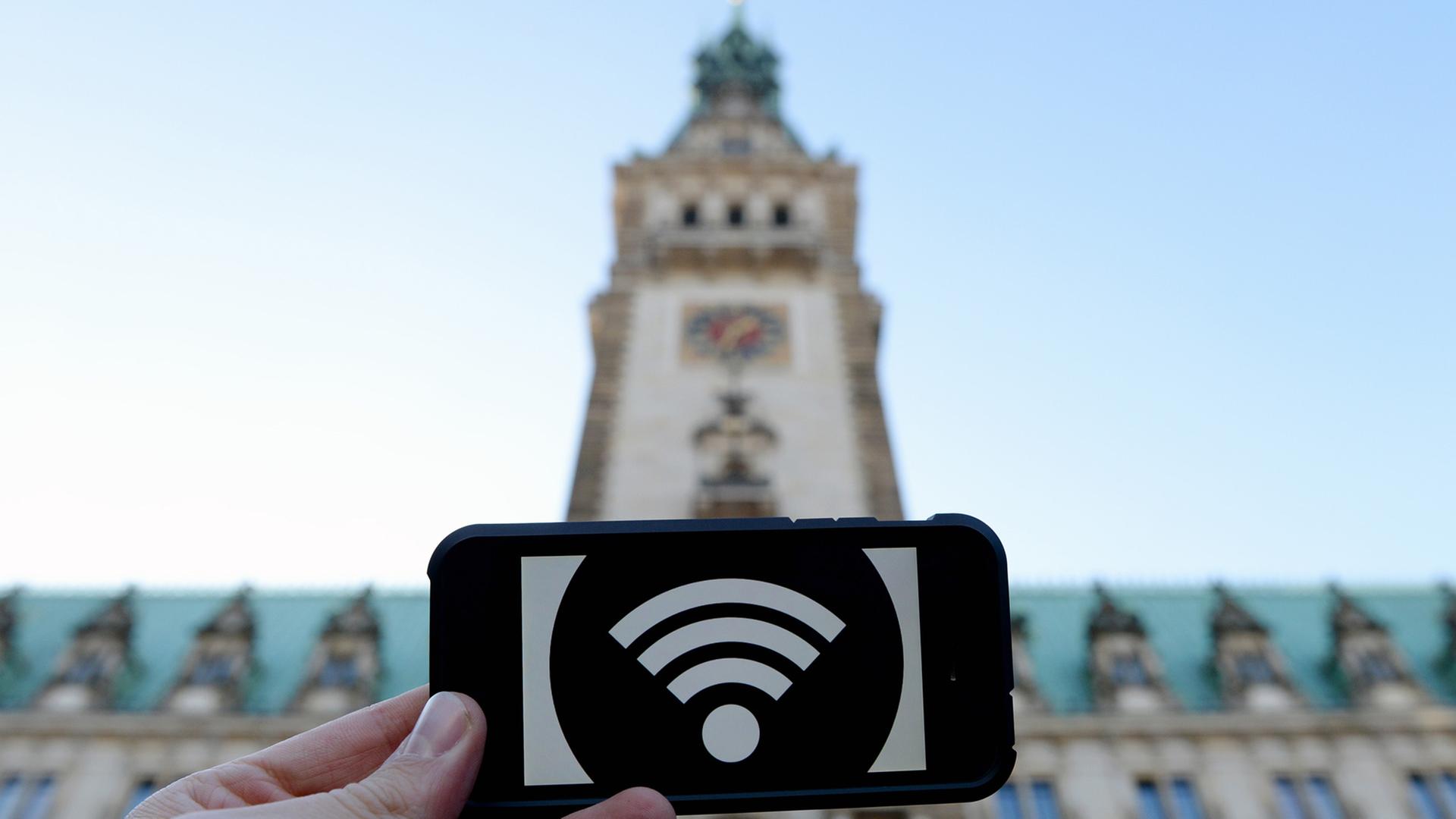 Eine Hand hält ein Schild mit einem WLAN-Symbol, im Hintergrund das Hamburger Rathaus.