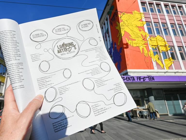 Eine Hand hält die Straßenzeitung "Asphalt", aufgeschlagen auf der Seite mit der kompletten Liste der ausstellenden Künstlerinnen und Künstler der Documenta 15, vor dem "ruruHaus".