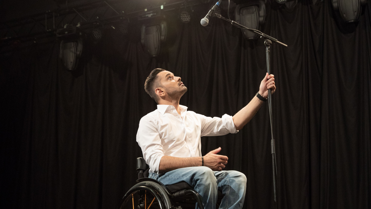 Tan  Caglar sitzt in seinem Rollstuhl auf der Bühne und blickt hoch auf ein Mikrofon. 