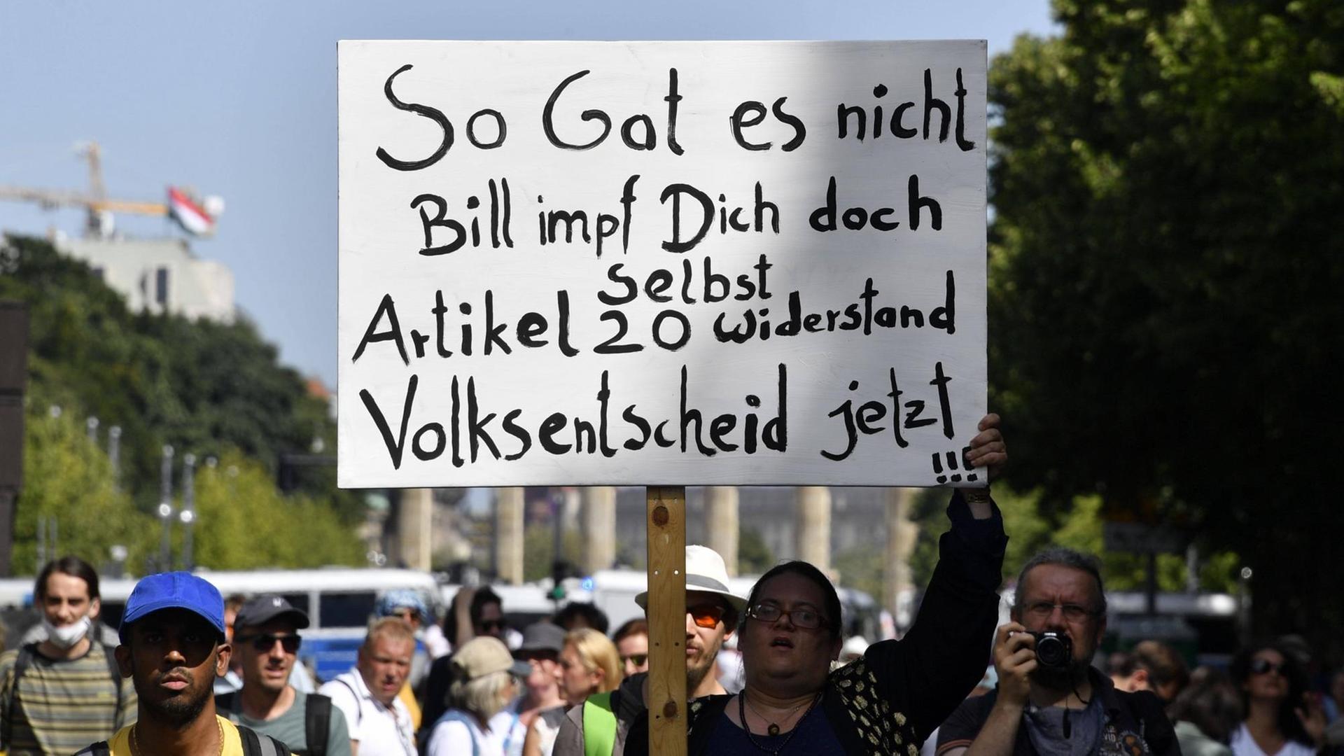Eine Demonstrantin hält ein Schild hoch mit der Aufschrift: "So Gates nicht. Bill impf Dich doch selbst. Artikel 20. Widerstand. Volksentscheid jetzt !!!"