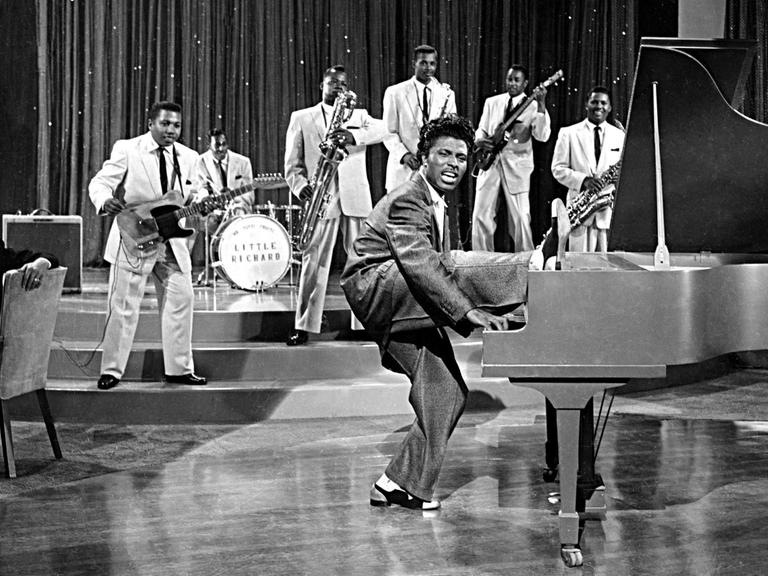 Eine historische Aufnahme in scharz-weiß zeigt den Musiker Little Richard am Klavier bei einer Fernsehaufzeichnung. Er hebt sein rechtes Bein. Im Hintergrund ist seine sechsköpfige Band zu sehen.