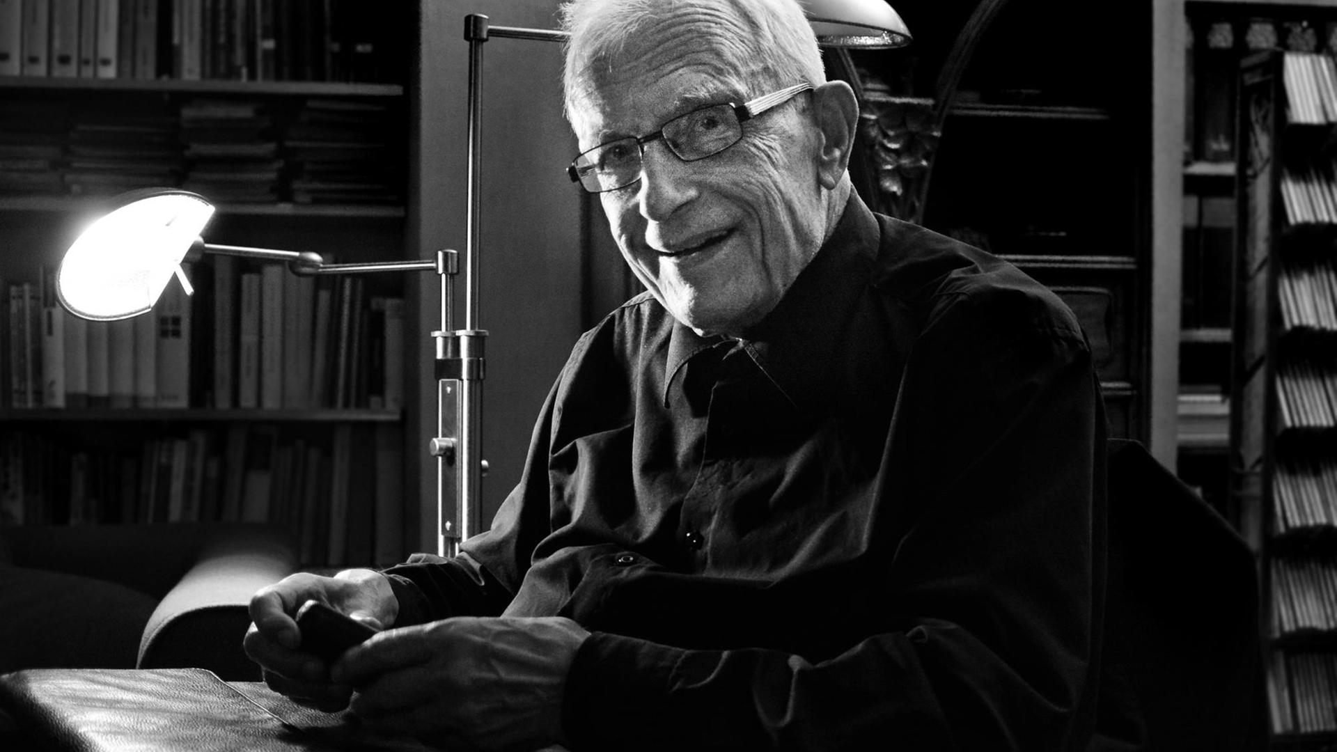 Hermann Schmitz, ein alter Herr mit weißem Haar und Brille, sitzt an seinem Schreibtisch und lächelt in die Kamera.